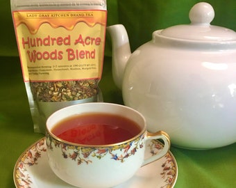 May include: A white teapot with a lid and a cup of tea on a saucer. The tea bag is labeled "Hundred Acre Woods Blend" and is from Lady Gray Kitchen Brand Tea. The tea bag lists the ingredients as Chamomile, Honeybush, Rooibos, Marigold Petals, and Vanilla Flavoring.