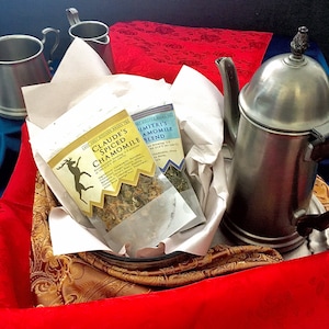 May include: A gift basket with a metal teapot, two small metal cups, and two tea packets. The tea packets are labeled "Claude's Spiced Chamomile" and "Dimitri's Chamomile Blend." The basket is lined with white paper and rests on a red and gold patterned fabric.