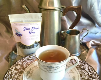 May include: A close-up of a cup of tea on a white saucer with gold trim. The tea is a light brown color. The cup is sitting on a tray with a silver teapot, creamer, and a package of Lady Gray Kitchen Brand Tea labeled "Glass Slipper Gray".