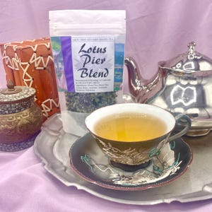 May include: A silver teapot and a teacup with a dragon design on a silver tray. The teacup is filled with a light yellow tea. A bag of Lady Gray Kitchen Brand Tea labeled "Lotus Pier Blend" is in the background.