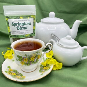 May include: A white teapot, sugar bowl, and teacup with a saucer are on a green surface. The teacup is filled with dark tea. A bag of Lady Gray Kitchen Brand Tea labeled "Springtime Blend" is in the background. The tea bag contains a blend of Rooibos, Chamomile, Lemongrass, Peppermint Leaf, Lemon Balm, and Marigold Petals.