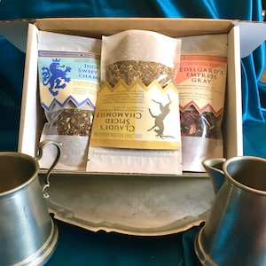 May include: A box of three different Lady Gray Kitchen Brand teas: Claude's Spiced Chamomile, Edelgard's Empress Gray, and Ingrid Sweet Chamomile. The teas are packaged in white paper bags with gold and black lettering. The box is open and the teas are visible. There are two silver mugs in the foreground.