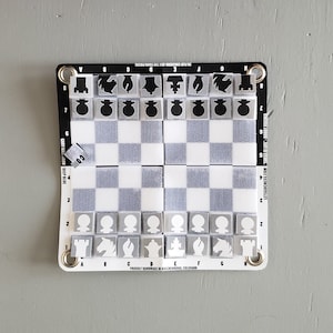 May include: A portable chess and checkers board with a white and gray checkered pattern. The board has a black border with the text "TO PLAY CHECKERS - USE THE PAWN PIECES" and "LETSCOCHESS.COM". The chess pieces are gray and white.