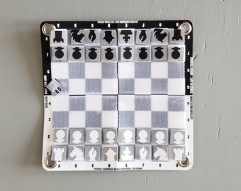 Op de afbeelding: Een draagbaar schaak- en dambord met een wit en grijs geblokt patroon. De zwarte rand draagt de tekst "TO PLAY CHECKERS - USE THE PAWN PIECES" en "LETSCOCHESS.COM". De schaakstukken zijn grijs en wit.