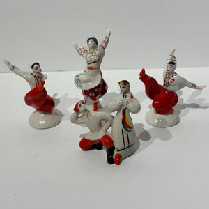 May include: A set of four porcelain figurines depicting dancers in traditional folk attire. The figures feature white shirts with red and black embroidered details, vibrant red trousers or skirts, and are posed in dynamic, expressive dance movements.