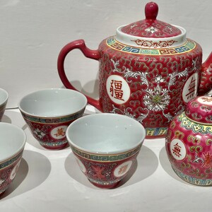 May include: A red and white Chinese tea set featuring a teapot, four cups, and two small jars. The set is decorated with gold accents and Chinese characters.