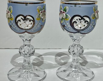 May include: Two clear glass shot glasses with a blue frosted finish. The glasses have a gold rim and are decorated with painted flowers and a gold and black design.