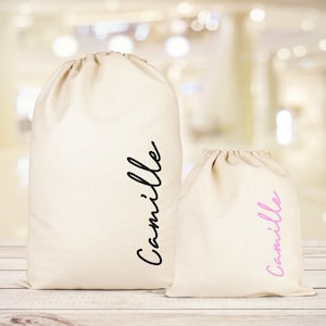 Puede incluir: Dos bolsas de tela blancas con el nombre "Camille" escrito en ellas. La bolsa m&aacute;s grande tiene el nombre en letras negras, y la bolsa m&aacute;s peque&ntilde;a tiene el nombre en letras rosas.