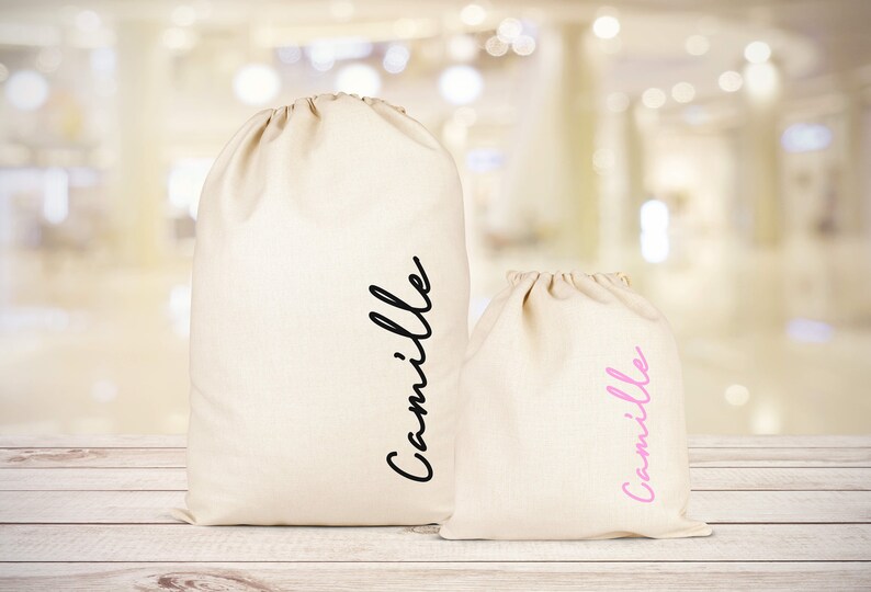 Puede incluir: Dos bolsas de tela blancas con el nombre "Camille" escrito en ellas. La bolsa m&aacute;s grande tiene el nombre en letras negras, y la bolsa m&aacute;s peque&ntilde;a tiene el nombre en letras rosas.