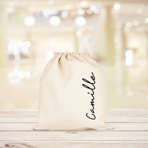 Puede incluir: Una bolsa de algod&oacute;n blanco con cord&oacute;n de ajuste con el nombre "Camille" escrito en cursiva negra en la parte delantera.
