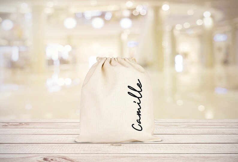 Puede incluir: Una bolsa de algod&oacute;n blanco con cord&oacute;n de ajuste con el nombre "Camille" escrito en cursiva negra en la parte delantera.