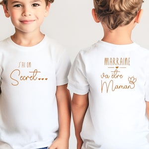 Könnte beinhalten: Zwei weiße T-Shirts mit braunem Text. Das erste Shirt sagt "J'ai un secret..." und das zweite Shirt sagt "Marraine va être Maman" mit einem kleinen Herz und einer Krone.