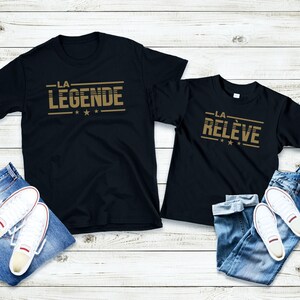 Peut inclure: Deux t-shirts noirs avec du texte dor&eacute;. Le plus grand t-shirt dit "LA LEGENDE" avec trois &eacute;toiles dor&eacute;es. Le plus petit t-shirt dit "LA RELEVE" avec trois &eacute;toiles dor&eacute;es. Les deux t-shirts sont dispos&eacute;s sur un fond en bois blanc avec un jean bleu et des baskets blanches.