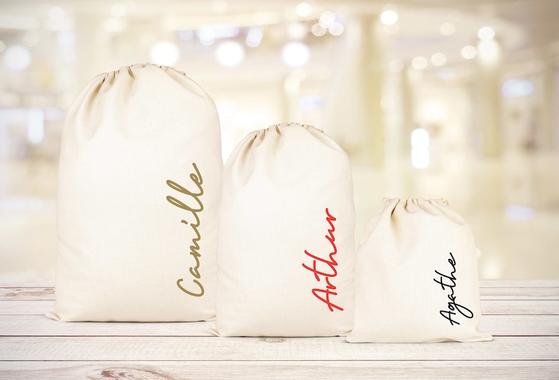 Puede incluir: Tres bolsas de tela blanca con cord&oacute;n de ajuste y nombres escritos en ellas. Las bolsas est&aacute;n ordenadas por tama&ntilde;o, la m&aacute;s grande a la izquierda y la m&aacute;s peque&ntilde;a a la derecha. Los nombres en las bolsas son Camille, Arthur y Agathe.