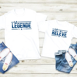 Peut inclure: Deux t-shirts blancs avec du texte bleu. Le plus grand t-shirt dit "LA LEGENDE" et le plus petit dit "LA RELEVE". Les deux t-shirts ont trois &eacute;toiles sous le texte.