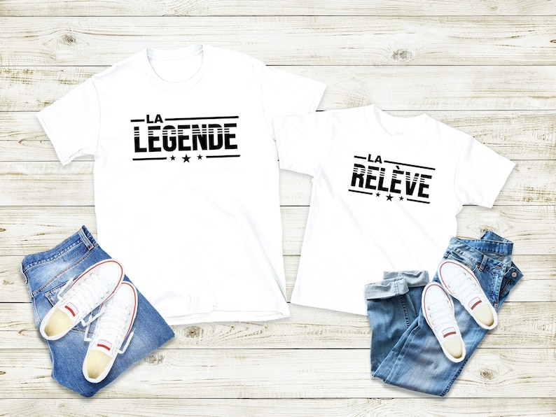 Peut inclure: Deux t-shirts blancs avec du texte noir. Le plus grand t-shirt dit "LA LEGENDE" avec trois &eacute;toiles. Le plus petit t-shirt dit "LA RELEVE" avec trois &eacute;toiles. Les deux t-shirts sont sur un fond en bois blanc avec un jean bleu et des baskets blanches.