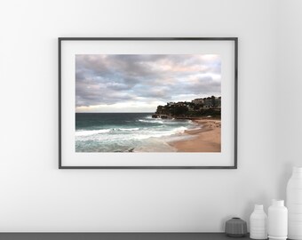 May include: A framed landscape photograph of a coastal scene featuring a sandy beach, turquoise ocean waves, and a rocky headland with buildings under a cloudy sky. The artwork is enclosed in a thin black frame and mounted on a plain wall.