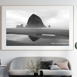 Könnte beinhalten: Ein schwarz-weiß Landschaftsfoto in einem hellen Holzrahmen, das einen großen Felsen im Meer an einem Sandstrand mit Spiegelung in einer Gezeitentümpel zeigt. Der Text Monika Hamilton Photography ist unten rechts auf dem Druck zu sehen.