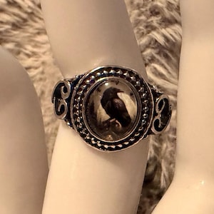 May include: A silver-toned ring featuring an oval-shaped centerpiece with a black and white graphic of a raven. The setting is surrounded by a beaded border and includes decorative scrollwork details on the sides of the band for a textured finish.