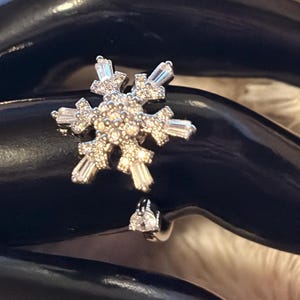 May include: A silver-toned ring featuring a prominent snowflake-shaped centerpiece adorned with numerous small, clear, sparkling stones. The snowflake design incorporates both round and baguette-cut stones, creating a detailed, symmetrical, and icy appearance.