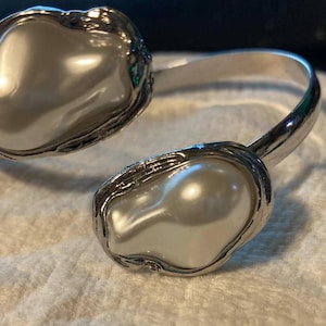 May include: A silver-toned cuff bracelet featuring two large, irregular-shaped faux pearls. The pearls are set in ornate, textured bezels. The bracelet has a simple, open design, with the pearls at each end.