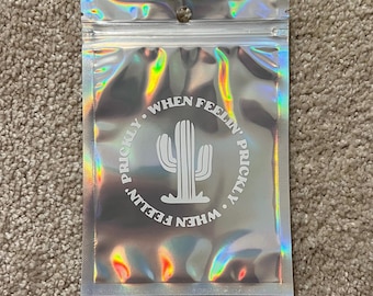 May include: A holographic zip-top bag with a white cactus design. The text "WHEN FEELIN' PRICKLY" is printed around the cactus. The bag is sealed and has a small hole near the top.