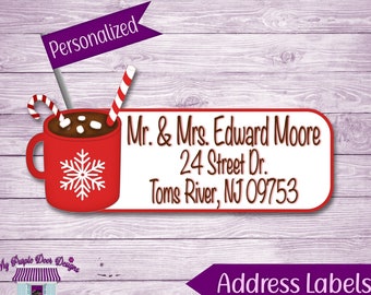 Op de afbeelding: Rood en wit adreslabel met een gepersonaliseerd bericht en een feestelijke illustratie van een rode mok met warme chocolademelk, marshmallows en snoepstokken. Het label bevat de tekst "Mr. & Mrs. Edward Moore, 24 Street Dr., Toms River, NJ 09753".