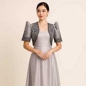 May include: A silver-gray midi dress with a square neckline and a matching bolero jacket. The jacket features short, structured puff sleeves and dark gray floral embroidery with sequin accents along the front edges and sleeve cuffs.