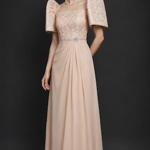 May include: A floor-length champagne-colored gown featuring a lace bodice with a square neckline and structured butterfly sleeves. The waist is accented with a central floral embellishment, transitioning into a flowing, pleated chiffon skirt.