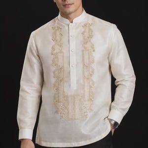 May include: A long-sleeved, off-white Barong Tagalog shirt featuring a mandarin collar, a partial button-down front, and intricate, symmetrical gold-toned embroidery panels running down the chest. The fabric has a subtle, semi-sheer sheen.