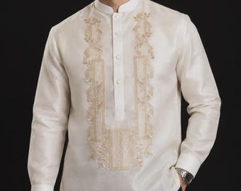 May include: A long-sleeved, off-white Barong Tagalog shirt featuring a mandarin collar, a partial button-down front, and intricate, symmetrical gold-toned embroidery panels running down the chest. The fabric has a subtle, semi-sheer sheen.