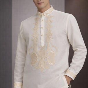 May include: Off-white long-sleeved shirt with intricate gold embroidery on the front, collar, and cuffs. The shirt has a mandarin collar and a button-down front. The garment is paired with black pants.