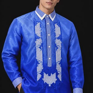 May include: A long-sleeved, royal blue barong tagalog shirt featuring a white pointed collar, white cuffs with a geometric pattern, and a central button placket. The front is decorated with white embroidered panels and intricate floral motifs.