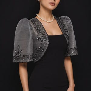 May include: A charcoal gray bolero jacket featuring short puff sleeves and intricate black floral embroidery with small clear sequins. The jacket has a scalloped edge along the front opening and sleeves, worn over a simple black sleeveless dress.