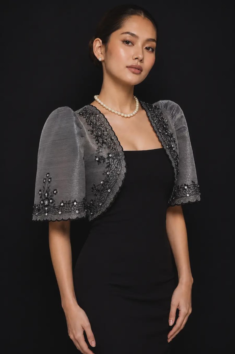 May include: A charcoal gray bolero jacket featuring short puff sleeves and intricate black floral embroidery with small clear sequins. The jacket has a scalloped edge along the front opening and sleeves, worn over a simple black sleeveless dress.