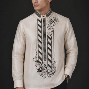 May include: A long-sleeved off-white barong tagalog shirt featuring a structured collar and a button-down front. The garment is decorated with black embroidered patterns, including diagonal stripes and intricate floral scrollwork on the chest.