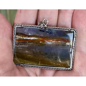 May include: A rectangular pendant featuring a polished, multi-colored stone with horizontal bands of deep blue, brown, and a thin streak of vibrant orange. The stone is encased in a silver-toned metal frame with a twisted rope-style border.