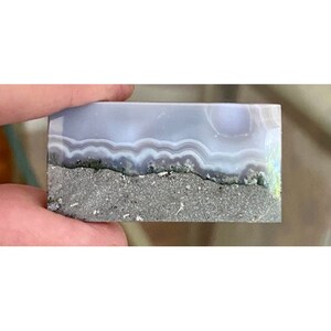 May include: A polished agate slab with a light blue and white banded pattern. The bottom of the slab is a rough, gray, and brown matrix.
