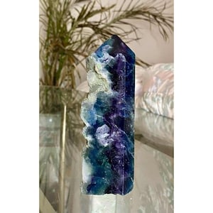 Könnte beinhalten: Ein polierter Fluorit-Kristallturm in sechseckiger Form mit einer spitzen Oberseite. Der Stein zeigt einen lebendigen Farbverlauf von tiefem Violett zu Blau-, Türkis- und Weißtönen, mit einer rauen, texturierten Kante an einer der Seiten.