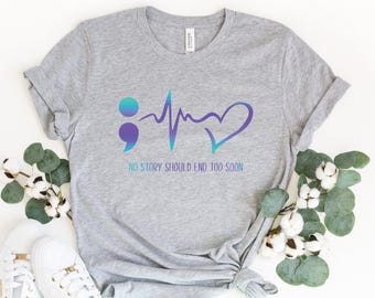 Może przedstawiać: Szary melanżowy t-shirt z krótkim rękawem z grafiką przedstawiającą średnik, linię tętna i serce w niebiesko-fioletowym gradiencie. Poniżej grafiki znajduje się napis NO STORY SHOULD END TOO SOON w pasującym niebiesko-fioletowym gradiencie.