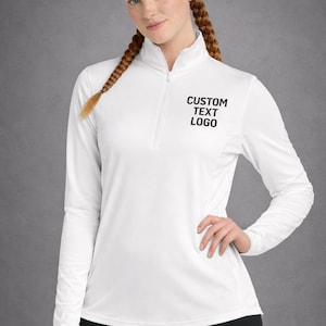 May include: A white long-sleeve athletic shirt with a quarter-zip collar. The shirt features the text "CUSTOM TEXT LOGO" in black. The model is wearing black pants.