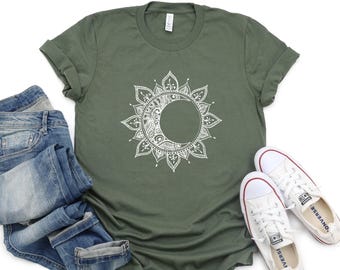 May include: An olive green short-sleeve t-shirt featuring a white graphic print of a crescent moon surrounded by intricate mandala-style floral patterns. The shirt is displayed alongside a pair of blue denim jeans and white canvas trainers.