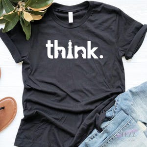 May include: A black short-sleeved t-shirt featuring the white text "think." printed across the chest. The letter "i" is replaced by a chess king piece, and the letter "n" is replaced by a chess rook piece. The shirt has a crew neckline.