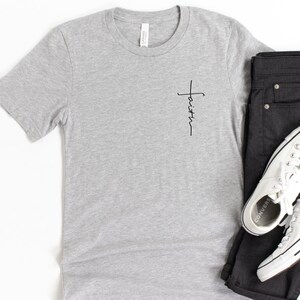 May include: A gray t-shirt with a small black cross and the word "faith" written vertically on the left side of the chest.