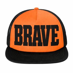 May include: A bright orange trucker hat featuring a black mesh back and a solid black flat brim. The front panel displays the word BRAVE in large, bold, black block lettering. The cap has a structured crown and a button on the top center.