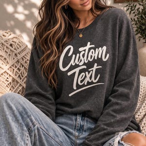 May include: A dark charcoal gray crewneck sweatshirt featuring the words Custom Text printed in a white script font across the chest. The garment has long sleeves with ribbed cuffs and a ribbed neckline, paired with light blue distressed denim jeans.