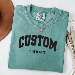 a turquoise t-shirt with the text "CUSTOM T-SHIRT" printed on it, along with a brown hat and a plant in the background.