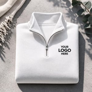 May include: A folded white quarter-zip sweatshirt with a silver zipper. The text "YOUR LOGO HERE" is printed in black on the front. The sweatshirt is on a gray surface with a white blanket and greenery in the background.