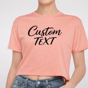 May include: A peach-colored short-sleeved crop top featuring the words Custom TEXT printed in black script and block lettering across the chest. The garment has a crew neckline and is paired with blue denim jeans.