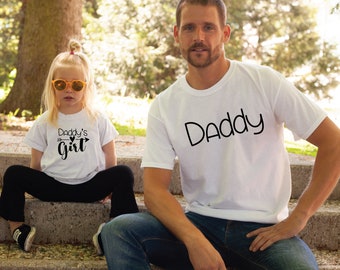 May include: A white t-shirt with the text "Daddy" in black. A white t-shirt with the text "Daddy's Girl" in black with a small heart and arrow graphic. The shirts are worn by a man and a girl.